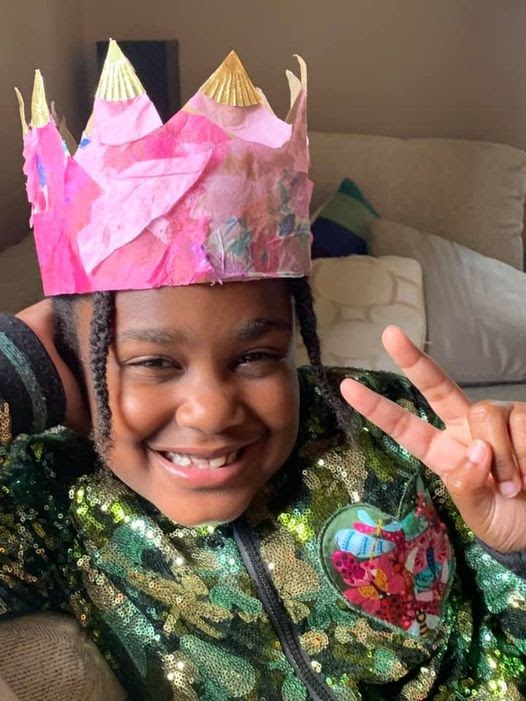 A happy girl wearing a pink paper crown and showing peace sign.