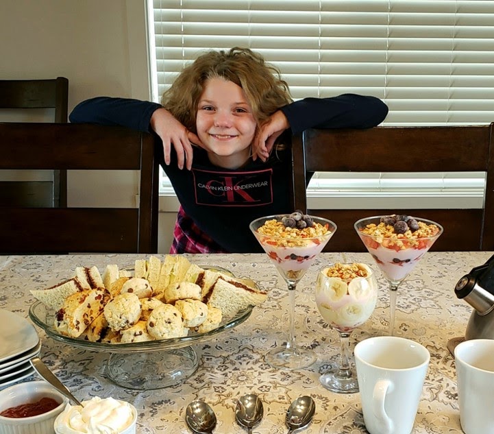 A girl happily standing in front of a bowl of scones and three glasses of parfait
